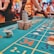 A casino scene with a roulette table features prominently, covered in green felt with colorful numbers and betting squares. Several stacks of poker chips are placed on different numbers, and the hands of several people are visible as they interact with the table. In the background, there are slot machines and more people, creating a vibrant and busy atmosphere.