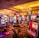 A vibrant casino interior filled with rows of colorful slot machines, each illuminated with bright lights and displays. The ceiling is adorned with elegant chandeliers and golden drapes, creating a luxurious atmosphere. The patterned carpet adds to the elaborate design of the space.