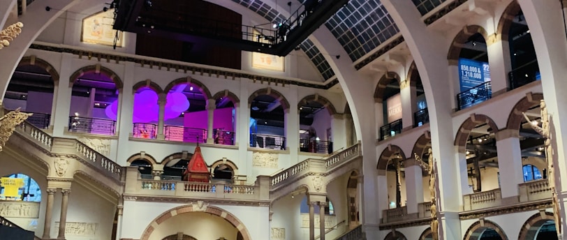 The interior of a large, ornately designed building with arched ceilings and intricate architectural details. The space features gaming machines lined up along the floor, under a high ceiling with a grid pattern. Lighting in purple and white tones enhances the ambiance.