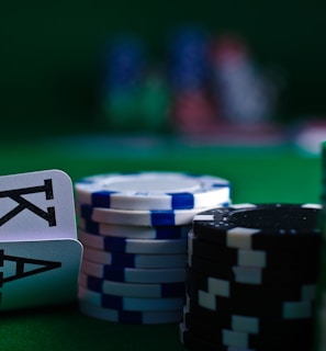 A casino scene featuring two playing cards, the ace and king of clubs, partially concealed by a player's hand. Nearby, there are stacks of multicolored poker chips on a green felt table, with blurred colorful background elements suggesting other chip stacks or cards.