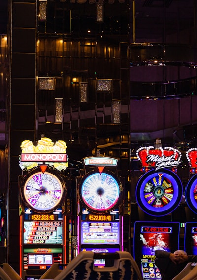 A lively casino scene featuring a row of bright, colorful slot machines with themes like Monopoly, Michael Jackson, and Lord of the Rings. The gaming machines display a variety of flashing lights and digital screens, creating an energetic and exciting atmosphere. Reflections of the lights are visible in glossy surfaces above the machines.